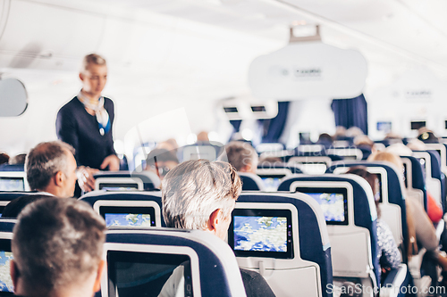 Image of Interior of airplane with passengers on seats and stewardess in uniform walking the aisle, serving people. Commercial economy flight service concept.