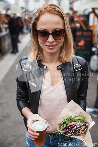 Image of Beautiful young woman holding delicious organic salmon vegetarian burger and homebrewed IPA beer on open air beer an burger urban street food festival in Ljubljana, Slovenia.