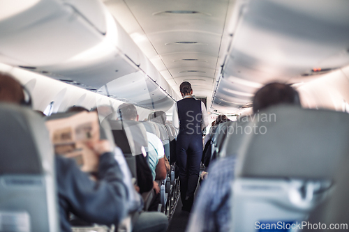 Image of Interior of airplane with passengers on seats and stewardess in uniform walking the aisle, serving people. Commercial economy flight service concept.