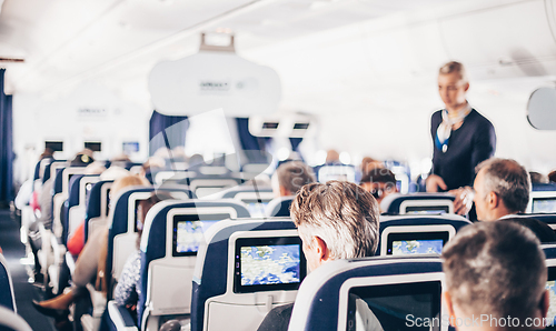 Image of Interior of airplane with passengers on seats and stewardess in uniform walking the aisle, serving people. Commercial economy flight service concept.