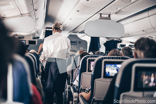 Image of Interior of airplane with passengers on seats and stewardess in uniform walking the aisle, serving people. Commercial economy flight service concept.