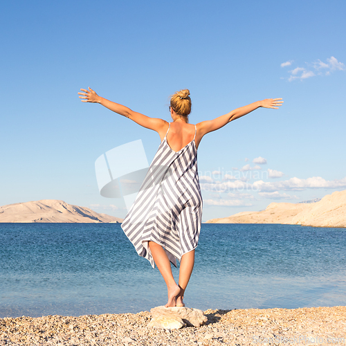 Image of Happy carefree woman rising arms, wearing beautiful striped summer dress enjoying late afternoon on white pabbled beach on Pag island, Croatia