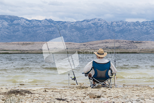 Image of Rear view of old unrecognizable fisherman wearing a straw hat, sitting in fishing chair with two fishing poles.