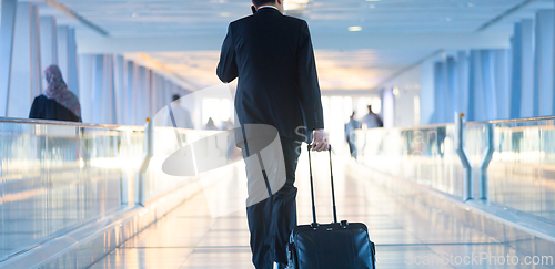 Image of Businessman walking and wheeling a trolley suitcase at the lobby, talking on a mobile phone. Business travel concept.