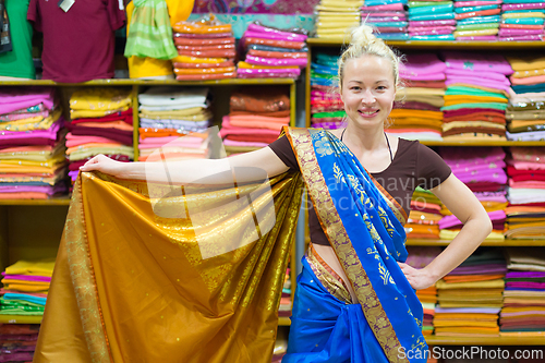 Image of Travel photo of beautiful blond Caucasian tourist woman trying to wear a traditional Indian saree in local saree shop as part of authentic travel experience.