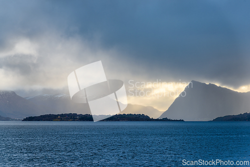 Image of Dramatic clouds gather over serene mountain landscape by the sea