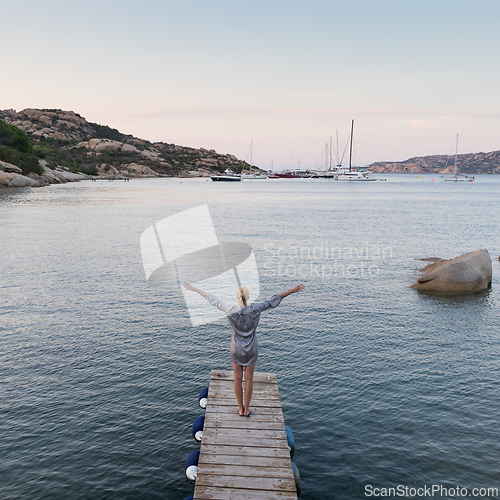 Image of Beautiful woman in luxury summer dress standing on wooden pier enjoying peaceful seascape at dusk. Vacation, resort and traveling.