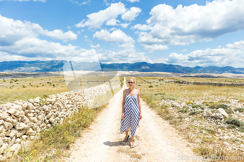 Image of Caucasian young woman in summer dress holding bouquet of lavender flowers while walking outdoor through dry rocky Mediterranean Croatian coast lanscape on Pag island in summertime