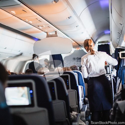 Image of Interior of airplane with passengers on seats and stewardess in uniform walking the aisle, serving people. Commercial economy flight service concept.