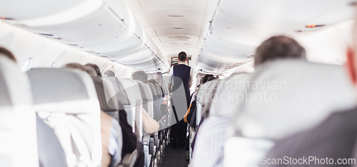 Image of Interior of airplane with passengers on seats and stewardess in uniform walking the aisle, serving people. Commercial economy flight service concept.
