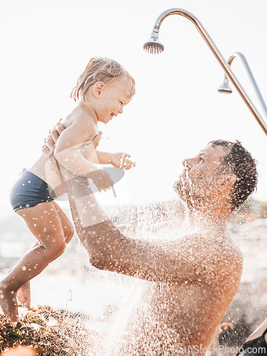 Image of Dad and baby under a tropical shower. dad and his cute infant boy playing on the beach under shower water splashes - Father taking care of cute sun in playful moment at summer family holiday.