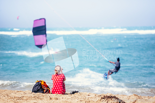 Image of Kiteboarder riding kite across the waves of Flag beach on vacations on Fuerteventura, Canary islands while her girlfriend watches him from the sandy beach, sunbathing.