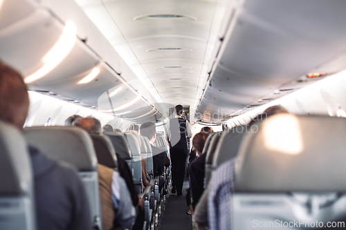 Image of Interior of airplane with passengers on seats and stewardess in uniform walking the aisle, serving people. Commercial economy flight service concept.