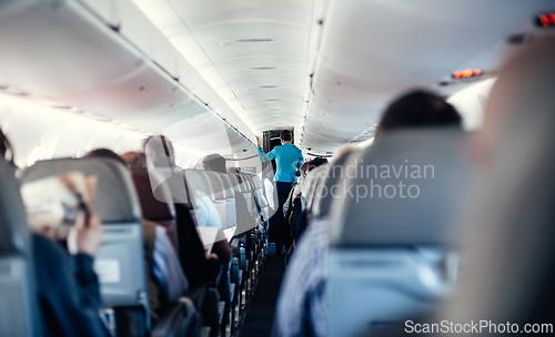 Image of Interior of airplane with passengers on seats and stewardess in uniform walking the aisle, serving people. Commercial economy flight service concept.