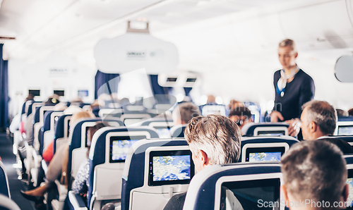 Image of Interior of airplane with passengers on seats and stewardess in uniform walking the aisle, serving people. Commercial economy flight service concept.