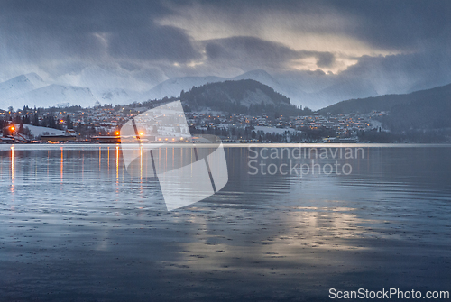 Image of Winter evening over a tranquil lake with mountains and lit build