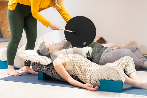Image of Group therapy session with Tibetan drum for group of women lying on wooden floor surrounded by copper bowls. Meditation and relaxation