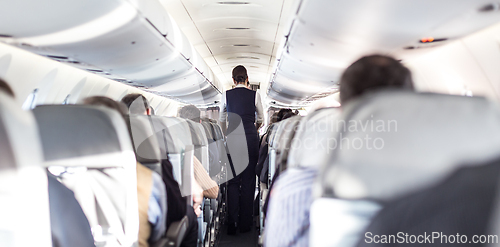 Image of Interior of airplane with passengers on seats and stewardess in uniform walking the aisle, serving people. Commercial economy flight service concept.