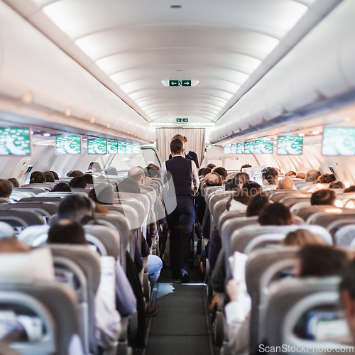 Image of Interior of airplane with passengers on seats and stewardess in uniform walking the aisle, serving people. Commercial economy flight service concept.