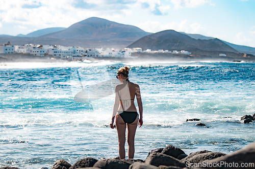Image of Female tourist at wild rocky beach and coastline of surf spot La Santa Lanzarote, Canary Islands, Spain. La Santa village and volcano mountain in background.