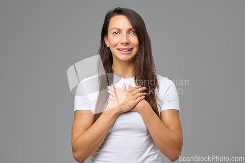 Image of Portrait of woman with hands on chest smiling softly in gesture of kindness and gratitude indoors
