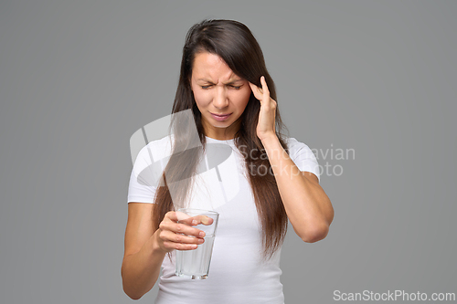 Image of Woman with headache holding glass of water expressing pain or discomfort against gray background