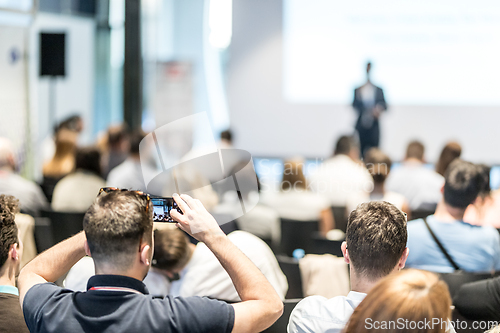Image of Male business speaker giving a talk at business conference event. Rear view of unrecognized participant in audience taking photo of presentation.