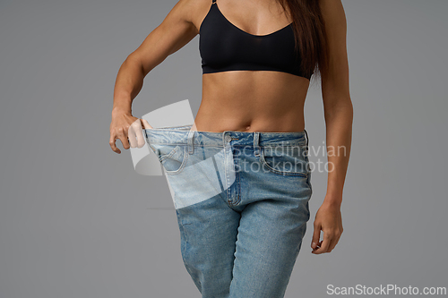 Image of Fit woman in black sports bra pulling loose jeans to show weight loss progress on gray background