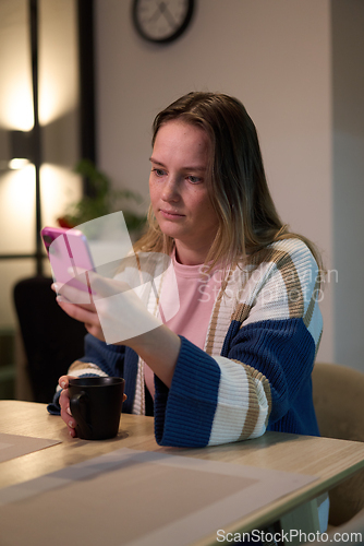 Image of Woman sitting at table looking at smartphone with cup in hand