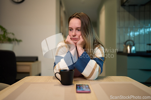 Image of Female resting head on hands at kitchen table with mug and smartphone