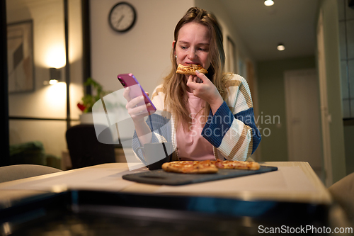 Image of Female eating pizza while using smartphone at kitchen table