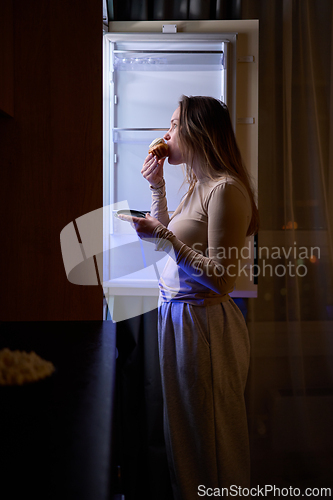 Image of Female eating muffin while standing in front of open refrigerator