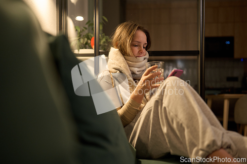Image of Female wrapped in scarf holding water and phone on sofa indoors