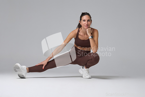 Image of Sporty woman stretching leg in side lunge pose on gray background in studio