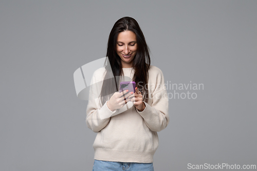 Image of Smiling woman looking at smartphone screen holding mobile device on grey background