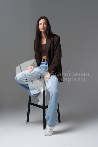 Image of Elegant woman with long dark hair wearing brown jacket and jeans sitting in studio