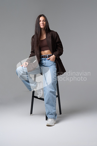 Image of Confident stylish woman posing on stool in brown jacket and light blue jeans
