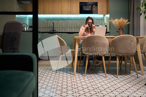 Image of Woman drinking from cup while sitting with laptop at kitchen table