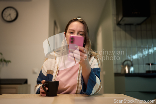 Image of Tired woman looking at phone screen with coffee mug in hand