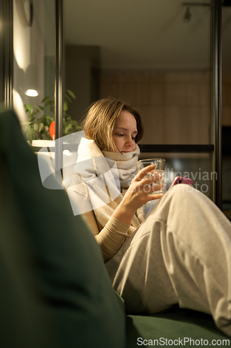 Image of Female sitting on sofa with scarf holding glass of water and phone