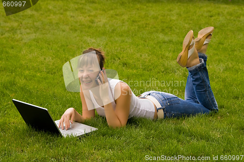 Image of Happy young woman with notebook and mobile phone on the grass