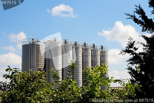 Image of Twelve high metal tower silos on chemical plant behind the trees
