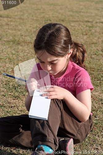 Image of Cute little girl with pigtails writes in the notepad sitting on the grass