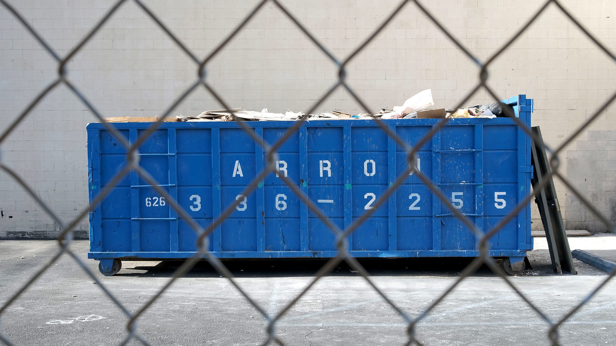 a big blue metal shipping container on a concrete floor in front of a concrete wall, the foreground is mesh metal screen