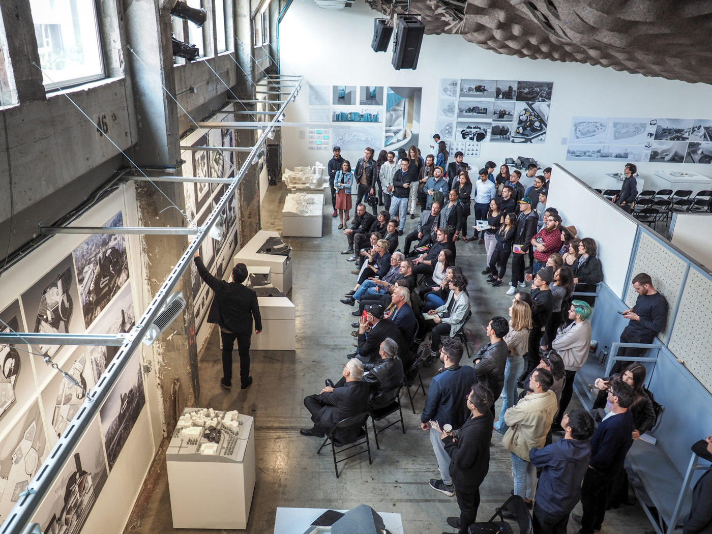 student in black outfit presenting his architecture project in front of a big crowd in a concrete and white building in SCI-Arc in downtown Los Angeles near Little Tokyo