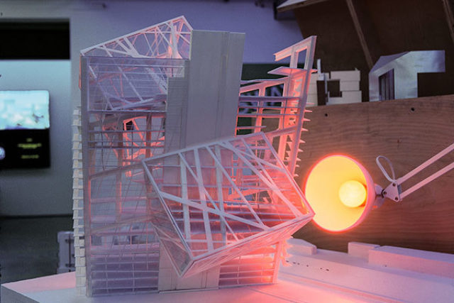 white deconstructed physical model of a building on top of a white model base lit by a warm desk lamp on the right