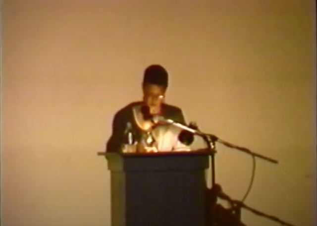 Michaele Pride, a Black female architect, lecturing at a podium in front of a white wall