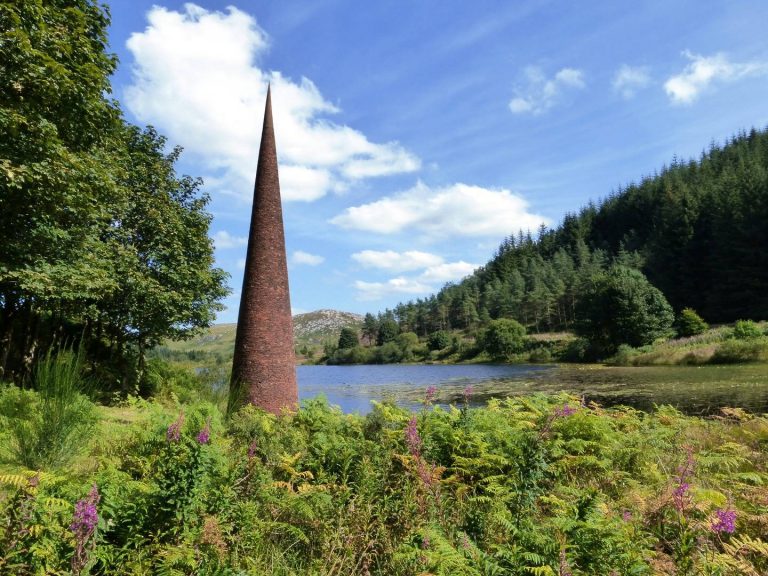 'The Eye' - a cone-shaped sculpture, 8 metres high and made from thousands of red tiles, designed by artist Colin Rose. Behind The Eye is Black Loch, with trees and the Galloway Hills in the far background. The sky is blue., Galloway & Southern Ayrshire Biosphere