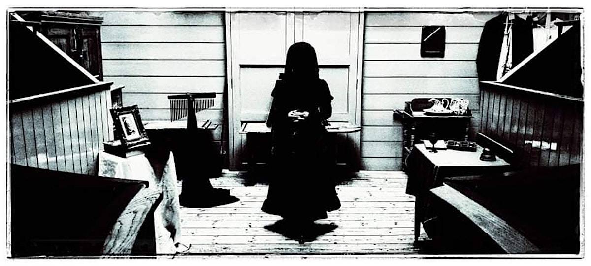 A black and white photograph showing a veiled woman in a black dress standing in a mysterious Victorian interior. We cannot see her face or what she is holding, but her form casts a shadow on the floor., Careena Fenton