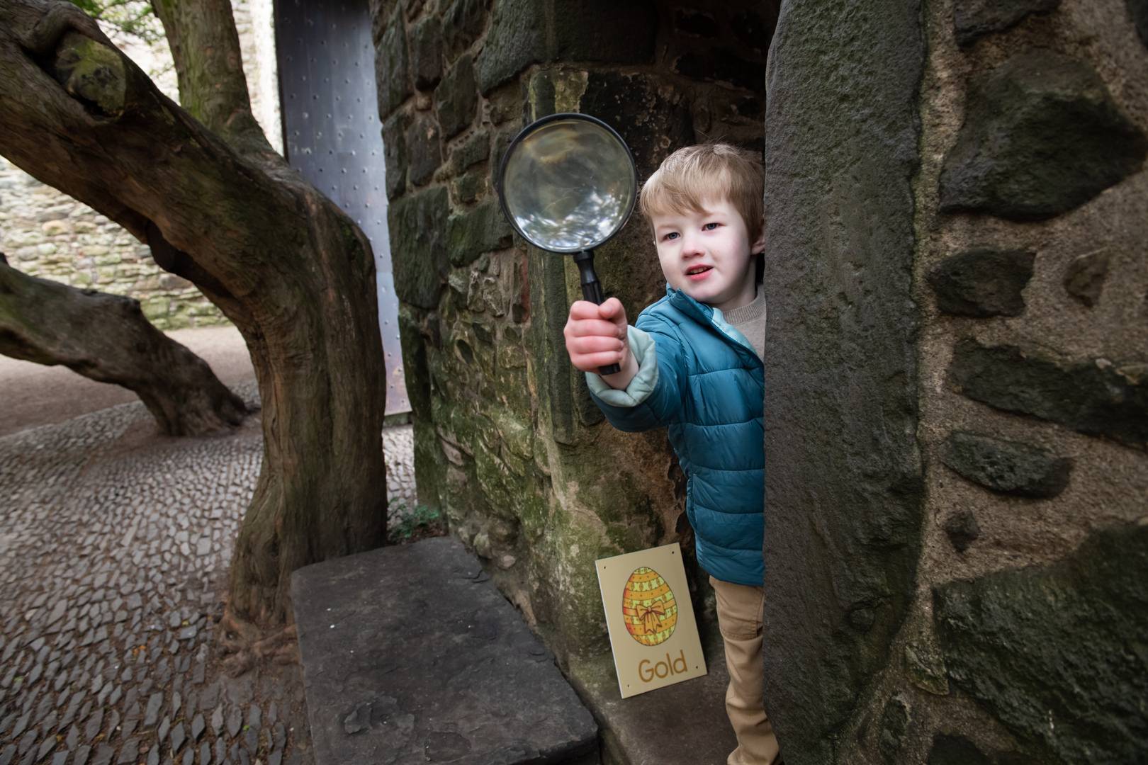 a little boy holds a magnifying glass as he peaks round a corner of a stone building